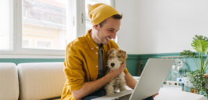 Young man wearing a winter hat sitting on a sofa holding his dog while looking at his laptop