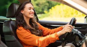 Young woman with long brown hair and a long sleeve orange shirt with both hands on the wheel driving her car